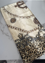 A folded beige silk-like fabric with a printed design featuring gold chain motifs, baroque scrollwork, and a leopard print section, displayed on a light gray marble surface.